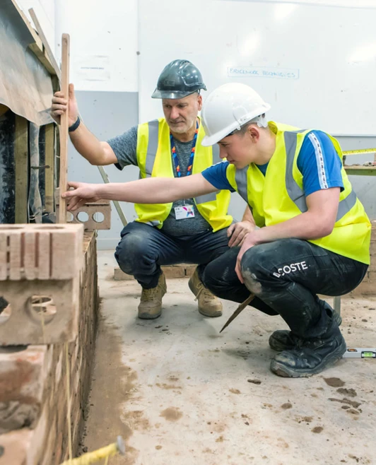 A lecturer instructing a student, both wearing high-visibility vests, as they assess a brickwork project in a hands-on college workshop. A lecturer instructing a student, both wearing high-visibility vests, as they assess a brickwork project in a hands-on college workshop.